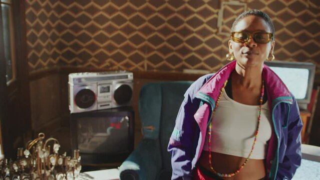Stylishly dressed Black girl in 80s shell suit moving her head to the beat and enjoying the music in old room, analog TVs with no signal screen and boombox in the background