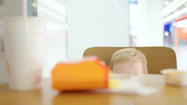 A cute blond toddler boy is hiding behind the table. A little child is shy and does not make contact with another person. Timid and bashful baby at a fast food restaurant