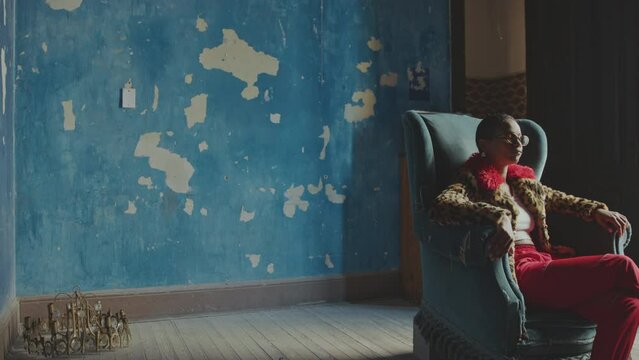Young eccentric girl in fur coat with leopard print and sunglasses sitting in old armchair and moving head to music beat in old vacated room with chipping paint on walls