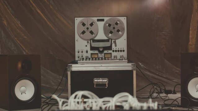 Dolly-out no people shot of working vintage reel to reel tape recorder, two speakers and tangled cables on floor in handmade music studio with polythene sheet wall