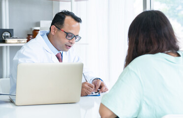 Fototapeta premium Woman patient visiting Indian male doctor at clinic office. Medical worker wear glasses giving healthcare consultation to Asian female patient, writes a prescription on a table in a hospital