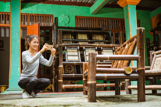 Asian girl squats using her phone camera to take a picture of a bamboo chair at a craft workshop