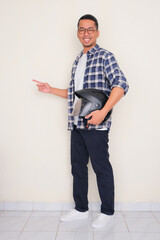 Full body portrait of a man smiling and pointing to the right side while holding motorcycle helmet