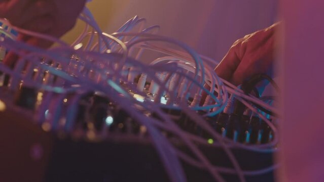 Unrecognizable DJ performing on modular synthesizer, plugging and unplugging audio cables on neon stage