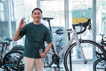 young asian man smiling with okay hand gesture while holding wrench with bicycle shop background