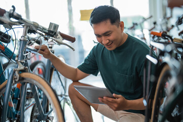 handsome young entrepreneur squatting to check a bicycle frame using a digital tablet at a bicycle shop - obrazy, fototapety, plakaty