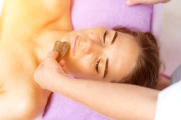 Fototapeta na wymiar Facial massage. Side view european woman getting massage with jade face roller gouache in spa salon