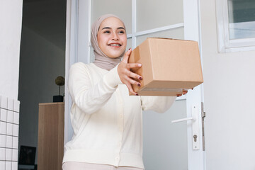 Cheerful Asian muslim woman receiving gift box delivery in front of the door at home