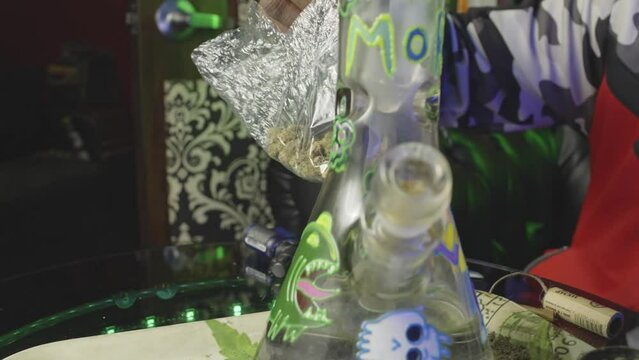 a black man lifts a bag of weed, removes a bud and smells it before breaking the marijuana up into the bowl of a water bong for smoking pot