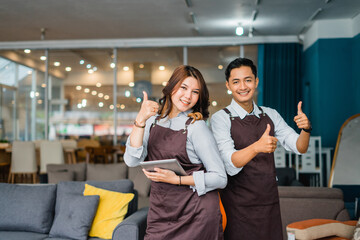 two shop assistants in aprons show thumbs up to the camera while standing in a furniture store