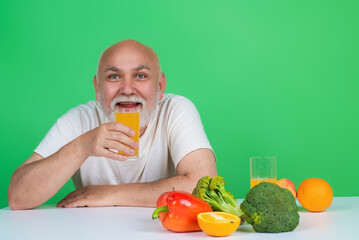 Senior drink orange juice near vegetables in studio. Elderly man hold glass of orange juice and vegetables. Orange juice and vegetables. Fruit and vegetables for aged people. Healthy lifestyle