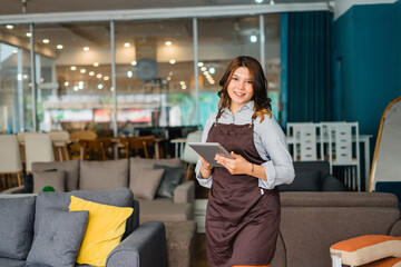 smiling female waitress in apron using a tablet digital standing in furniture store department