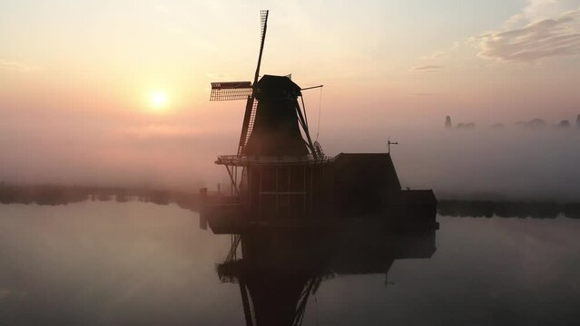 The drone is flying around a windmill with the sun rising revealing behind the windmill and lots of mist in de Zaanse Schans The Netherlands Aerial Footage 4K