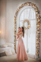 Fototapeta premium A little beautiful girl in a vintage dress stands with her mother near the panoramic window. Mom holds the baby. Family. Holiday. Mom and daughter look out the window. Mom gently hugs her daughter.
