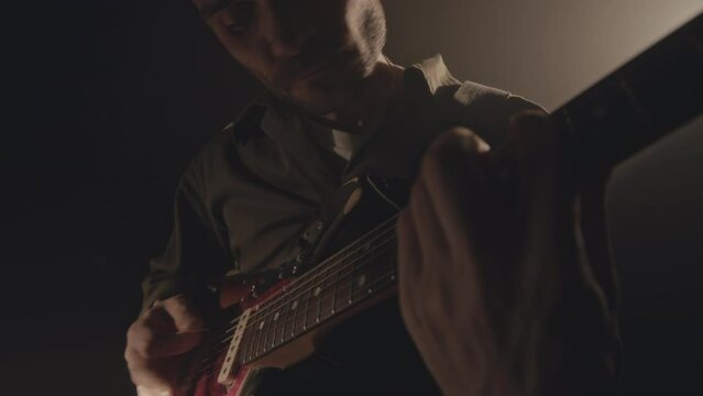 Tilt up shot of skilled young Caucasian male musician playing electric guitar masterly in dark room