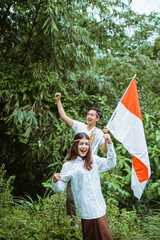 Fototapeta premium a beautiful woman in white shirt with ribbon at her haed standing with a handsome man grab the indonesian flag at the background