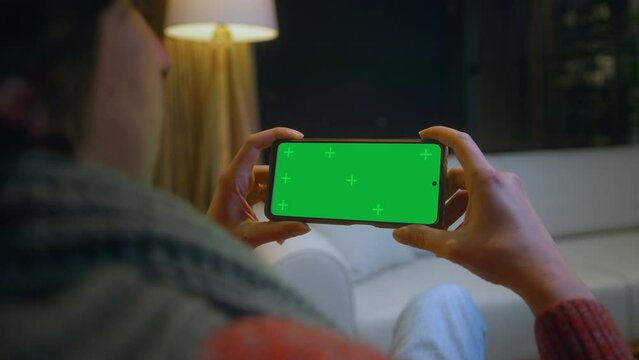 Young man watching video on his smartphone with green screen mock up display, which he holds horizontally. Male sitting on sofa relaxing at home. Close-up over the shoulder shot