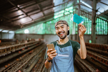 Smiling Asian businessman showing a bankbook and holding a phone standing at a chicken farm