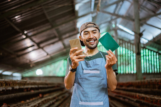 Smiling Asian businessman works with cell phone and passbook at chicken farm