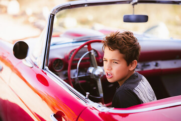 Young boy sitting in red classic car pretending driving - obrazy, fototapety, plakaty
