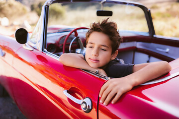Young boy sitting in red classic car day dreaming