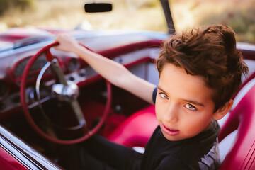 Young boy sitting in red classic car pretending driving - obrazy, fototapety, plakaty