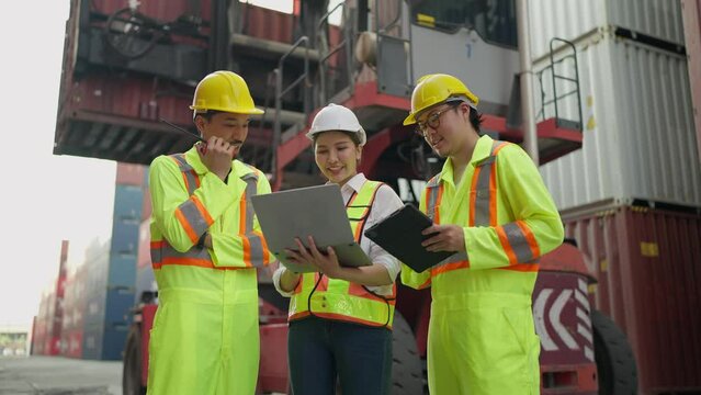 Hardworking team of workers in an industrial warehouse, wearing safety helmets working on laptop and tablet focused discusstion and talking together at cargo container yard. Teamwork concept