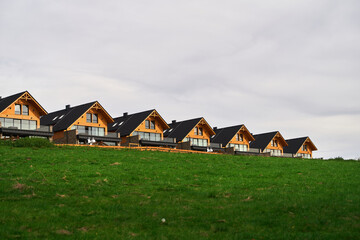 Fototapeta na wymiar A row of modern wooden houses nestled on a lush green hill under a cloudy sky offering a blend of nature and architecture.