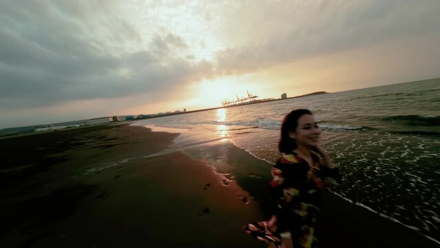 drone follow shot, young girl runs away in sunset sandy beach The sunshine is reflected on the beach with the sunset clouds behind with harbor.