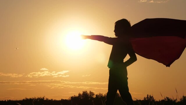 Silhouette of a superhero with his arm stretched forward against the background of the sun during sunset. A child in a superhero cape stands on top of a mountain, his cape fluttering in the wind.