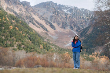 Serene journey, Asian woman in a blue jacket stands by the lake, surrounded by fall foliage. An elegant portrait of nature's beauty and traveler's happiness.