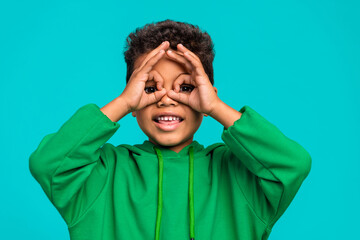 Photo of cheerful carefree cute boy dressed trendy clothes showing gesture looking through hole isolated on cyan color background