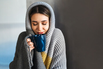 Young woman at home in winter wrapped herself in a warm cozy blanket, holding a cup of hot drink