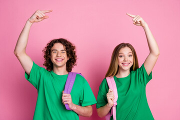 Portrait of two optimistic school children wear green t shirt and backpacks point fingers above...