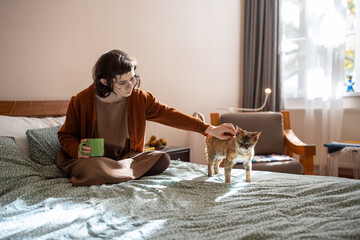 Girl petting Devon Rex cat sitting in bed at home drinking coffee at home. Woman stroking domestic pet. Spending morning time with animal starting new day. Life of lonely introverted girl with cat 