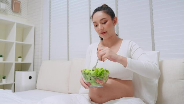 Happy pregnant Asian woman eating salad vegetable on the bed in bedroom at home. Food while pregnancy concept
