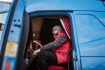 Portrait of confident professional truck driver sitting in truck cabin, looking at camera, smiling.