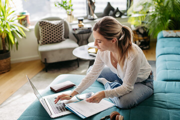 Woman paying bills online, sitting in living room, working on notebook. Online banking, internetbanking at home throught website, app. - obrazy, fototapety, plakaty