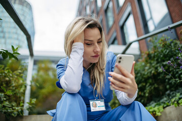 Young female doctor feeling overwhelmed at work, sitting on stairs, looking at phone. Healthcare...