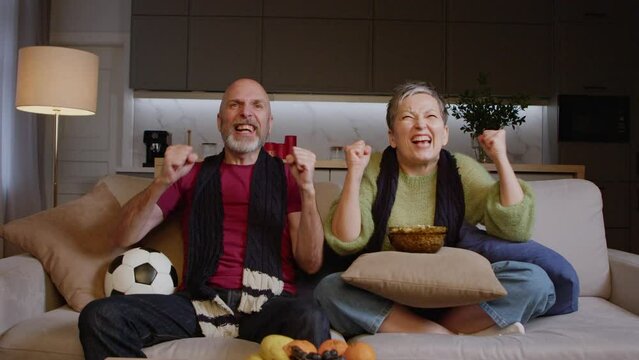 Excited good looking mature man with soccer ball and pretty middle aged woman with popcorn watching football match on tv, emotionally celebrating goal with fist pump gesture indoors.