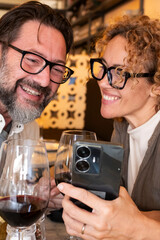 Happy couple smiling and talking in a restaurant looking at cell phone - Married couple enjoying lunch break at cafe bar - People and technology concept
