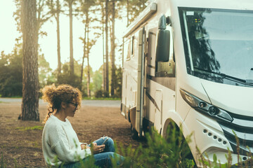 Serene woman enjoying nature sitting outside a modern big motorhome camper van with high trees and...