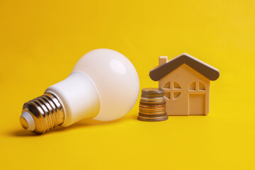 White lightbulb, stacked coins and a house miniature over yellow background. Concept of saving...