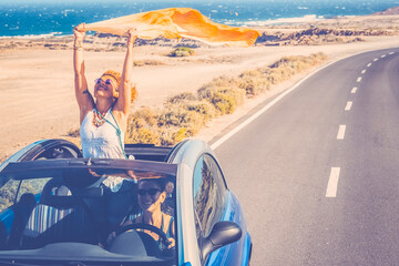 Happy women traveler enjoy road trip with a convertible car. One woman outside the roof have fun. Scenic desert destination. Car driving. Excited couple of friends in a vehicle on travel. Adventure - obrazy, fototapety, plakaty
