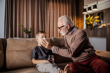 An ill boy is sitting at home on sofa while his granddad is blowing his nose.