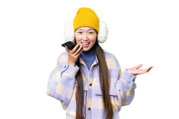 Young Asian woman wearing winter muffs over isolated chroma key background keeping a conversation with the mobile phone with someone