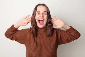 Cheerful funny surprised brown haired adult woman wearing brown sweater posing isolated over gray background raised her arms screaming with amazement exclaiming