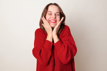 Attractive cheerful funny Caucasian young adult woman wearing red jumper touching her cheeks smiling toothily with shy expression posing isolated over gray background