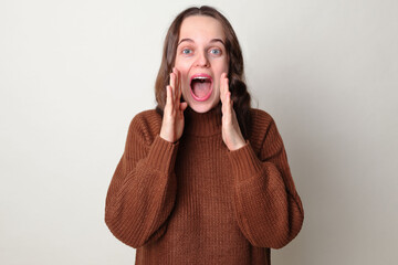 Amazed Caucasian woman wearing brown jumper standing isolated over gray background screaming with hands near mouth making advertisement yelling loud announcing good important news