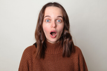 Shocked astonished Caucasian woman wearing brown jumper standing with open mouth and big eyes...